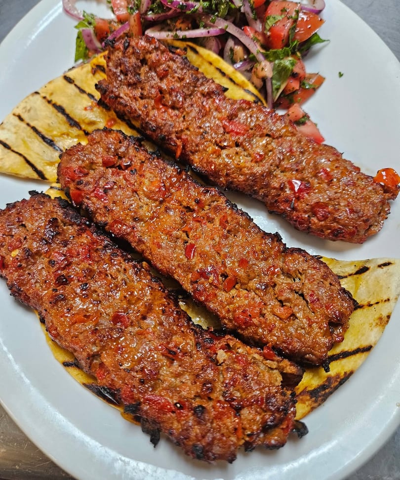 Three grilled Adana kebabs with red peppers on flatbread, served with a side salad.