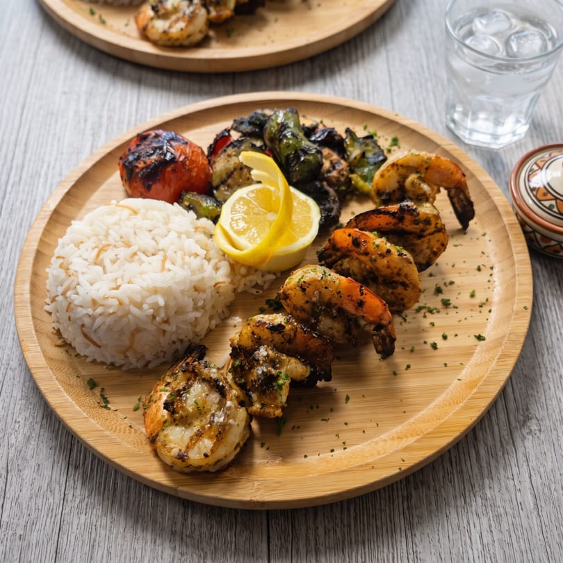Grilled shrimp, rice, charred tomatoes, and peppers on a bamboo plate with a lemon wedge.